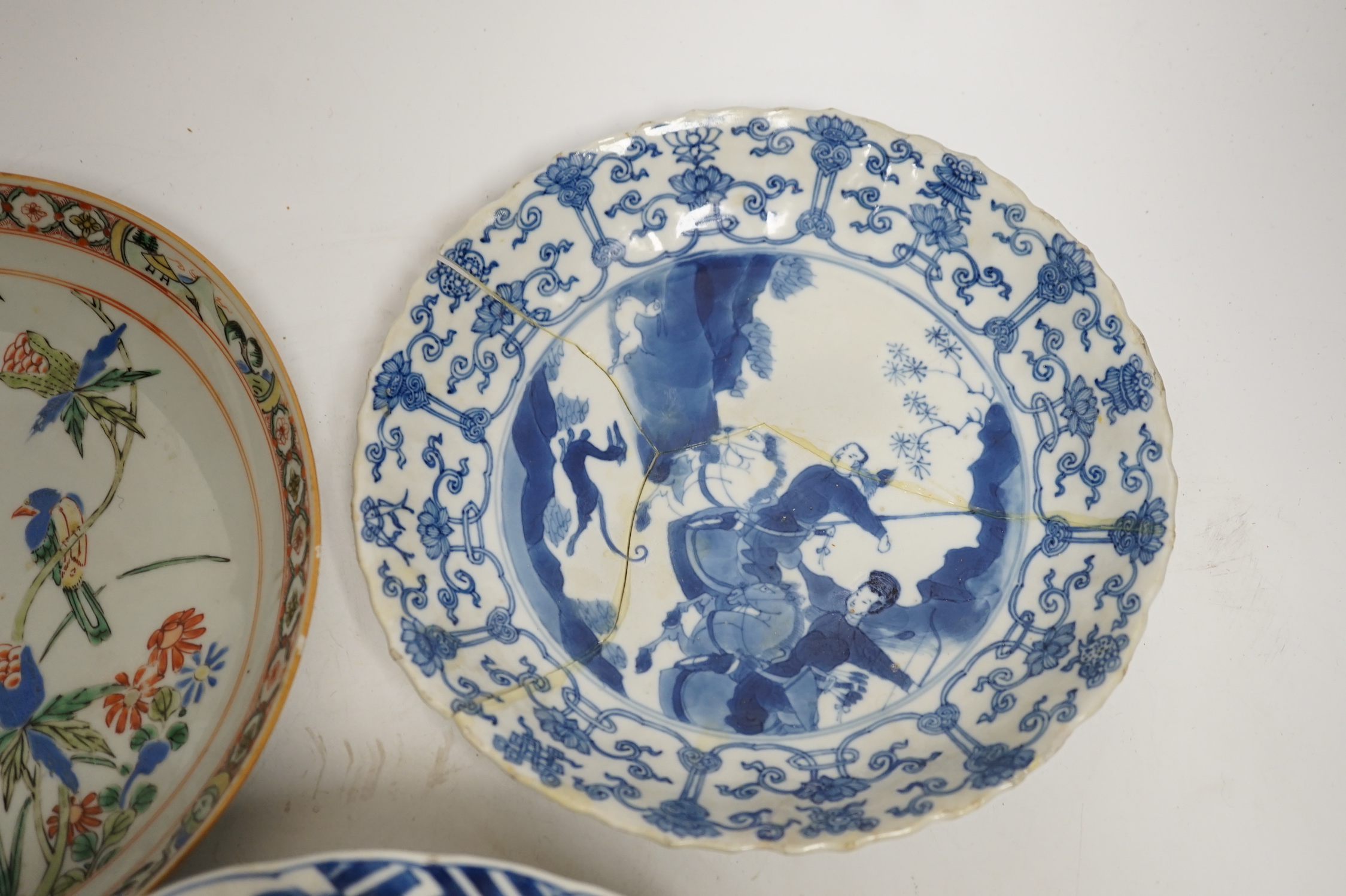 A Chinese blue and white ‘warriors’ bowl, Kangxi mark and period, a similar ‘hunting’ saucer dish, and a famille verte saucer dish, all Kangxi period (3), largest 27cm in diameter